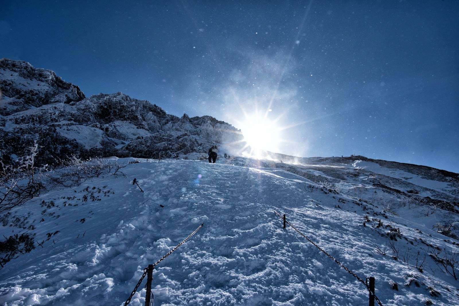 雪に覆われた山の稜線から雪煙が舞い上がり、青い空に太陽が輝く冬山の風景