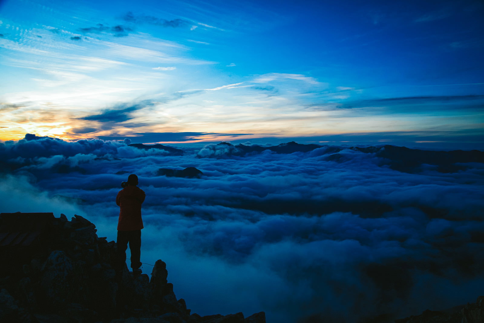 朝焼けの空と雲海を背景に、山頂で立つ登山者のシルエット
