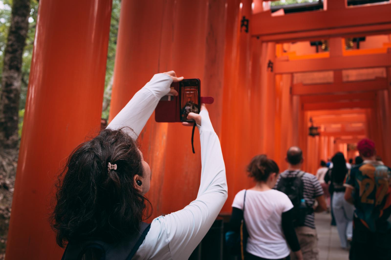 伏見稲荷大社の朱塗り千本鳥居でスマートフォンを掲げて撮影する白い服の外国人女性観光客