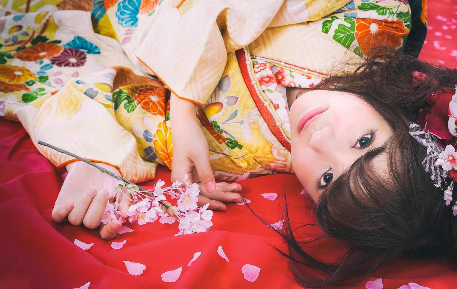 赤い毛氈に寝転がり桜の花びらに包まれた振袖姿の女性［モデル：河村友歌］