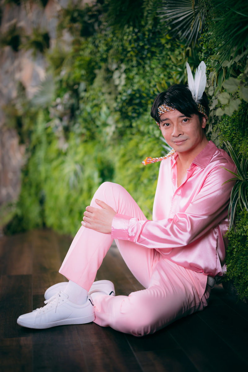 ピンクスーツにヘッドドレスをつけた男性が観葉植物の前で宣材撮影をしている［モデル：金子周平］