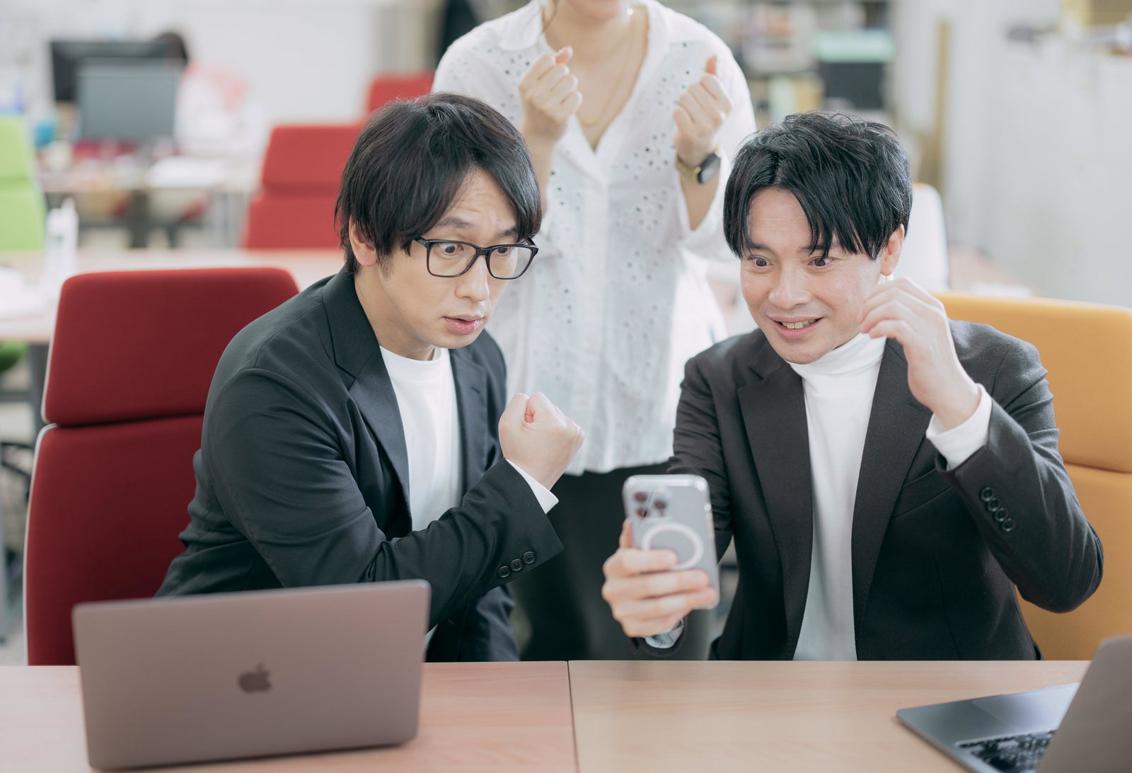 スマートフォンを持ってガッツポーズする男性社員と応援する女性社員［モデル：大川竜弥 金子周平］