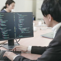 サブモニターのコード画面を見るエンジニア男性の写真