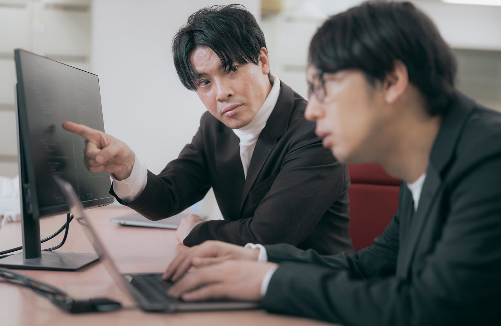 オフィスの机でパソコンモニターを指差しながら指示する男性と聞き手［モデル：金子周平 大川竜弥］