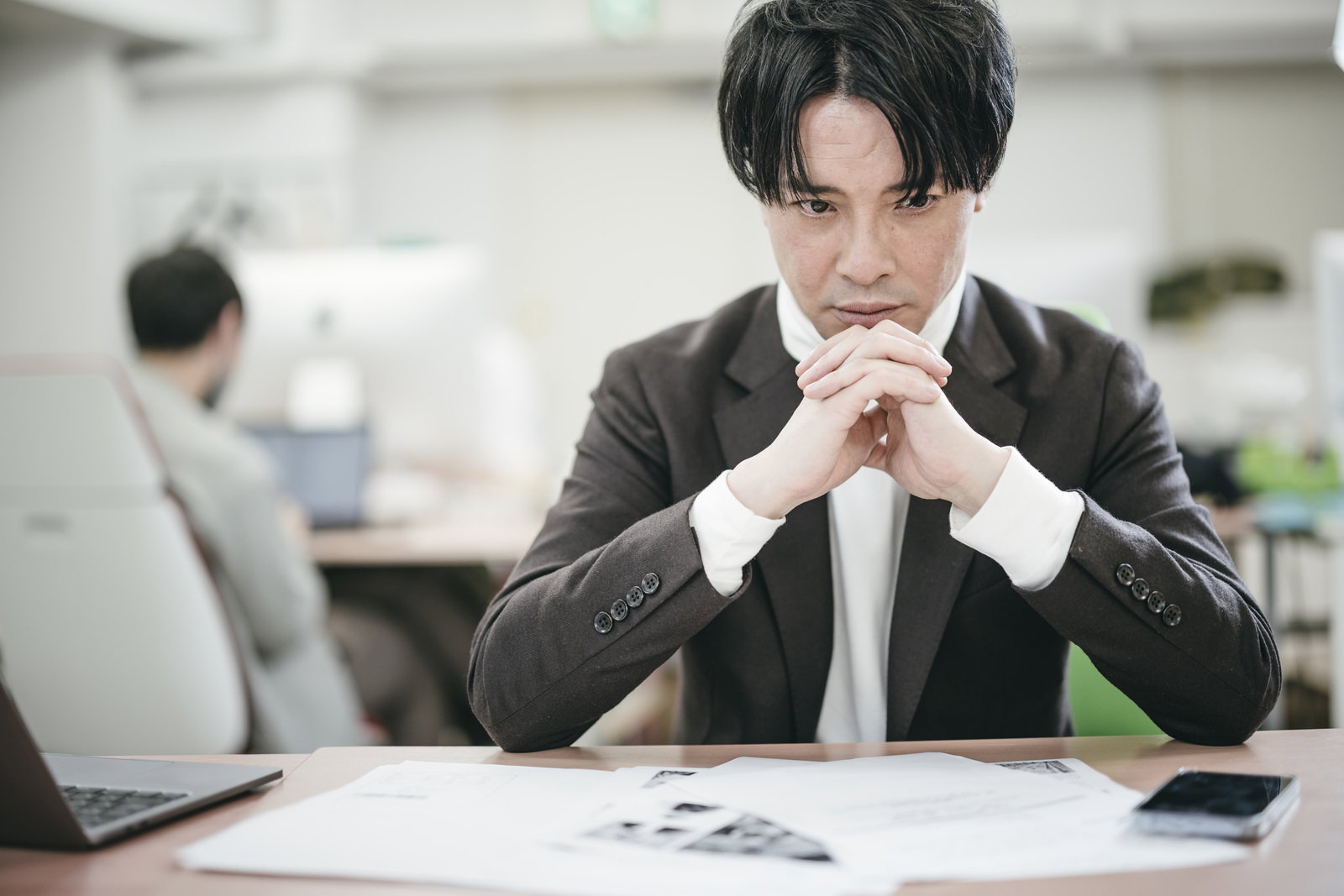 オフィスの机で書類を前に考え込むスーツ姿の男性社長［モデル：金子周平］