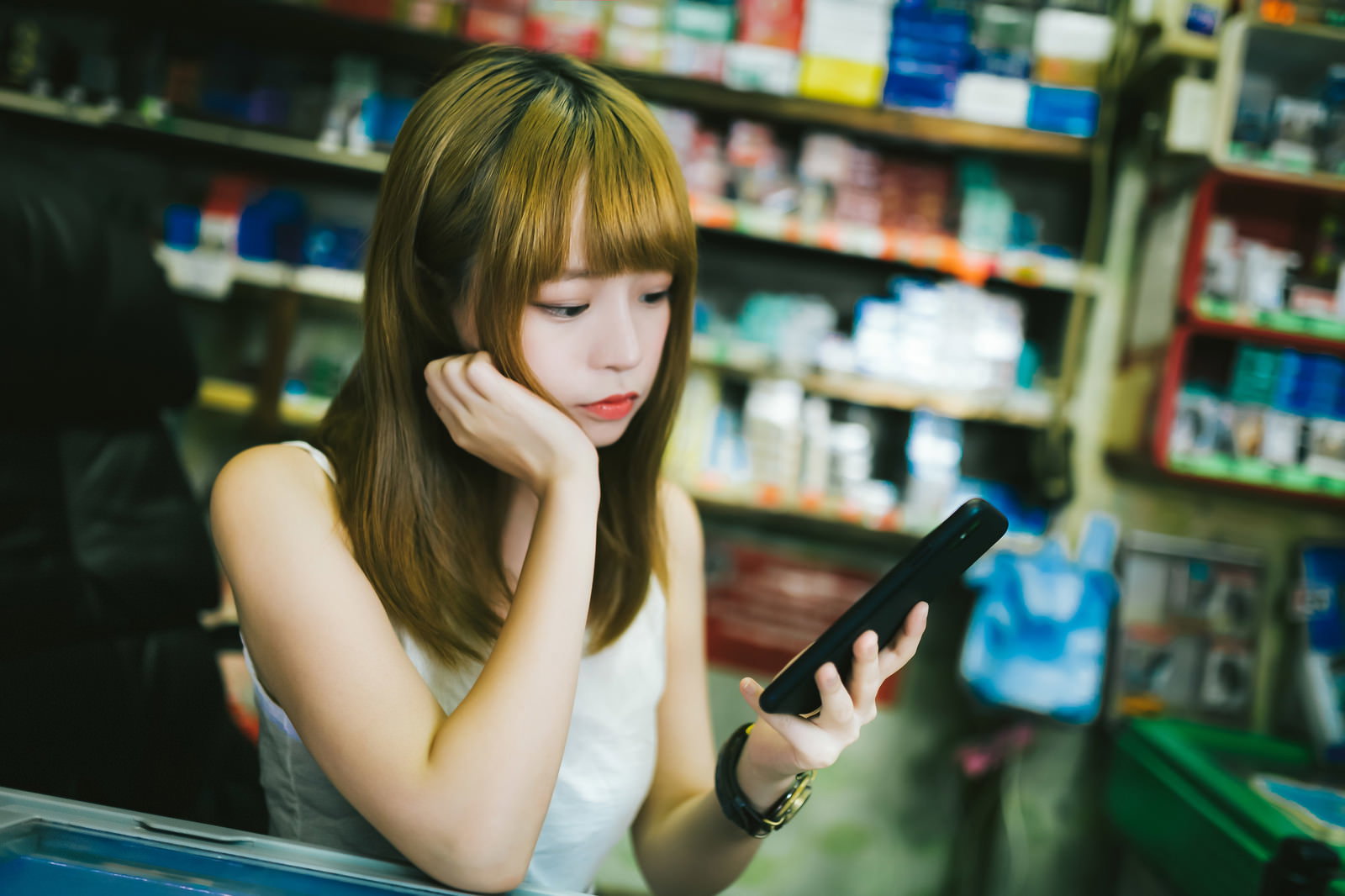 店番をしながら頬杖をついてスマホを見るプリン髪の女性［モデル：まめち］