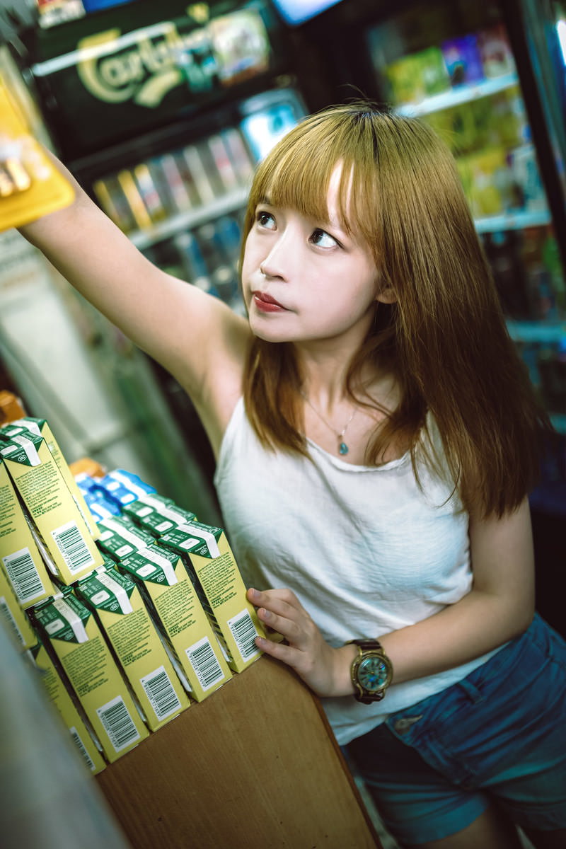 プリン髪の女性が背伸びをして棚上の商品を取ろうとしている［モデル：まみち］