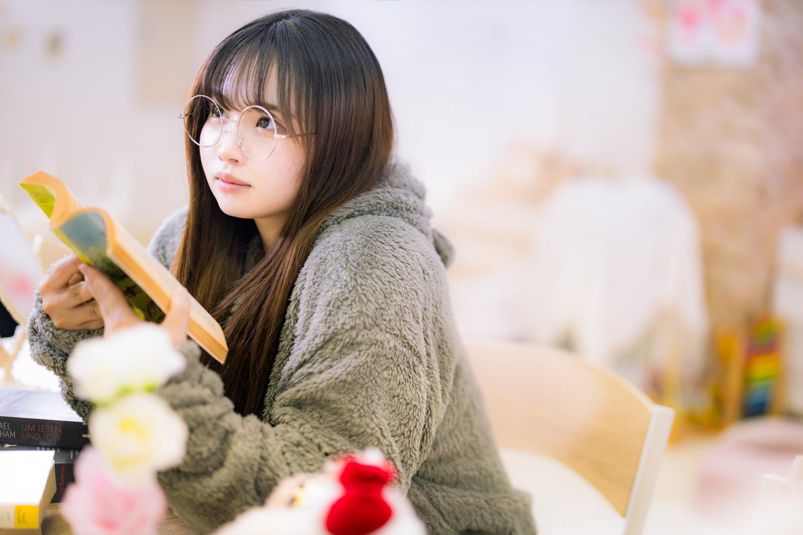 本を手に持ったままふと視線を上げる、フリースを着た丸メガネの女性［モデル：カナ］
