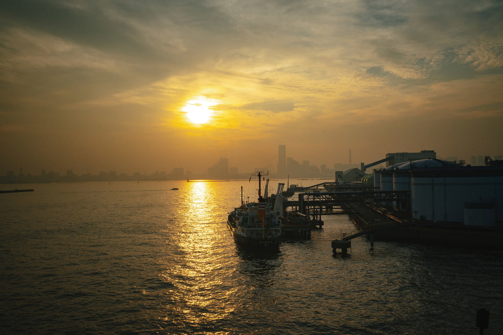 工業地帯の港で夕日が海面に反射し、船や建物がシルエットになっている風景