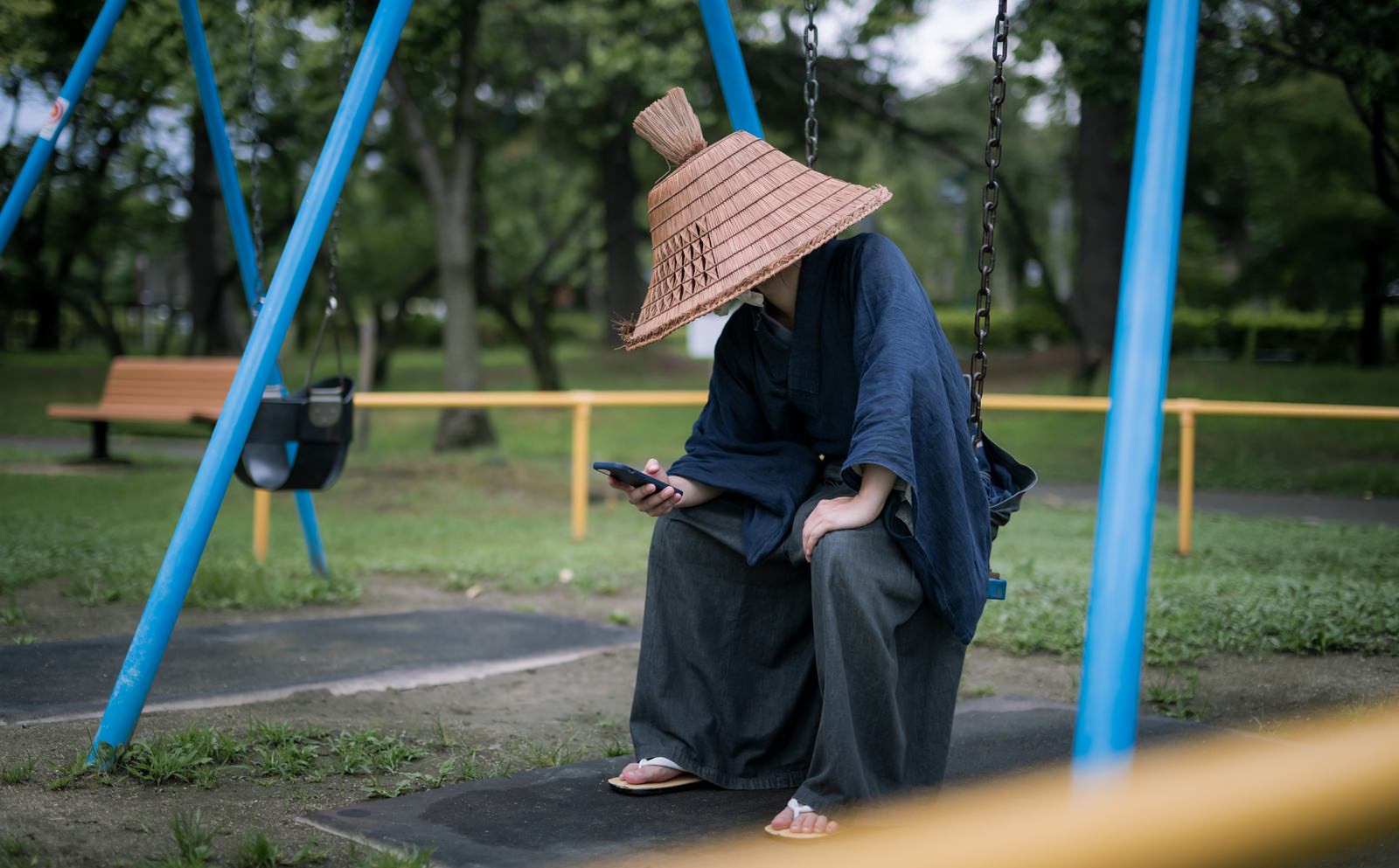網代笠を被った男性が公園のブランコに座ってスマートフォンを見ている［モデル：まーこ］