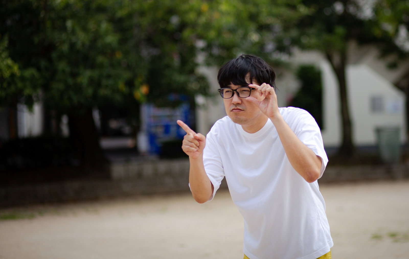 白いTシャツを着た眼鏡の男性が屋外で片手で眼鏡を押さえながら指差し確認をしている［モデル：真崎真幸］