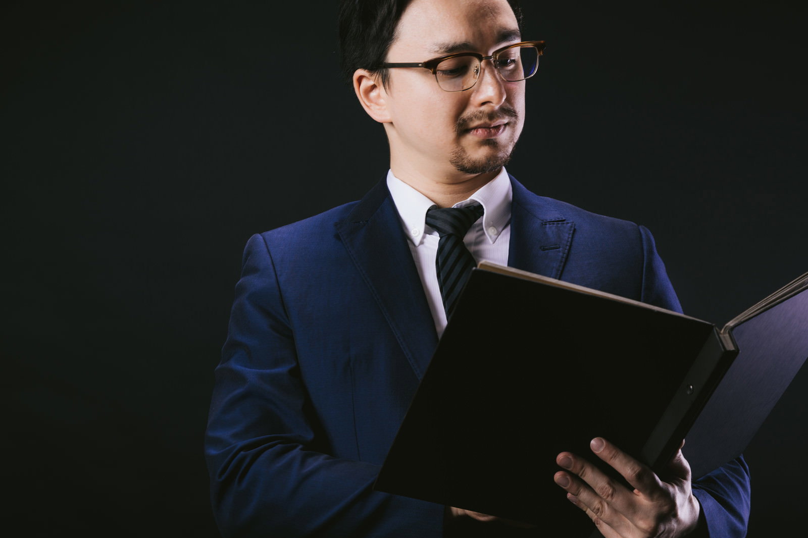 スーツを着た眼鏡の男性が書類ファイルを手に持って確認している［モデル：Max_Ezaki］