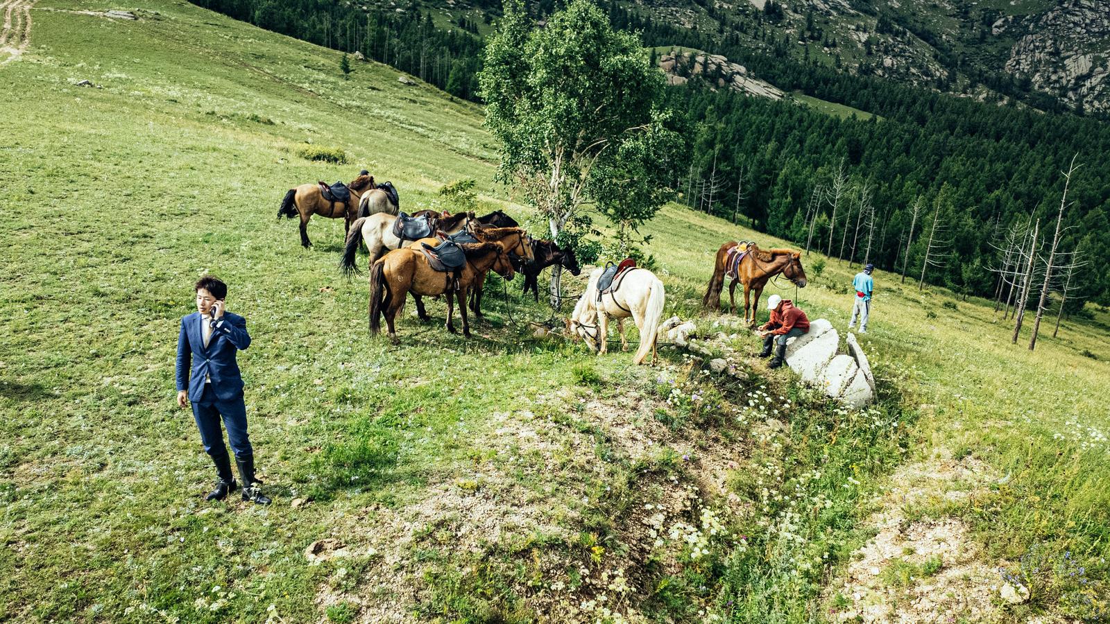モンゴルの草原でスーツ姿の男性が馬に乗りながら通話している［モデル：野崎繁裕］