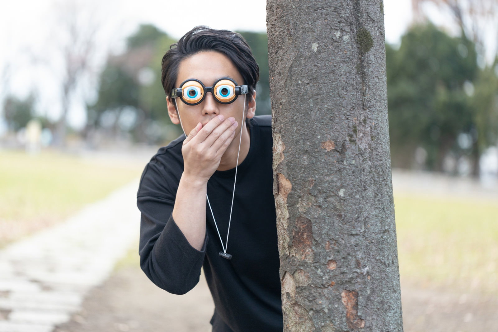 木陰から顔を出して目を丸くして驚いている眼鏡をかけた男性［モデル：大川竜弥］