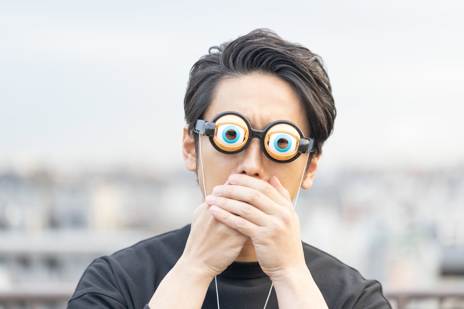 大きな丸メガネをかけた男性が手で口元を覆い驚いた表情を見せている［モデル：大川竜弥］