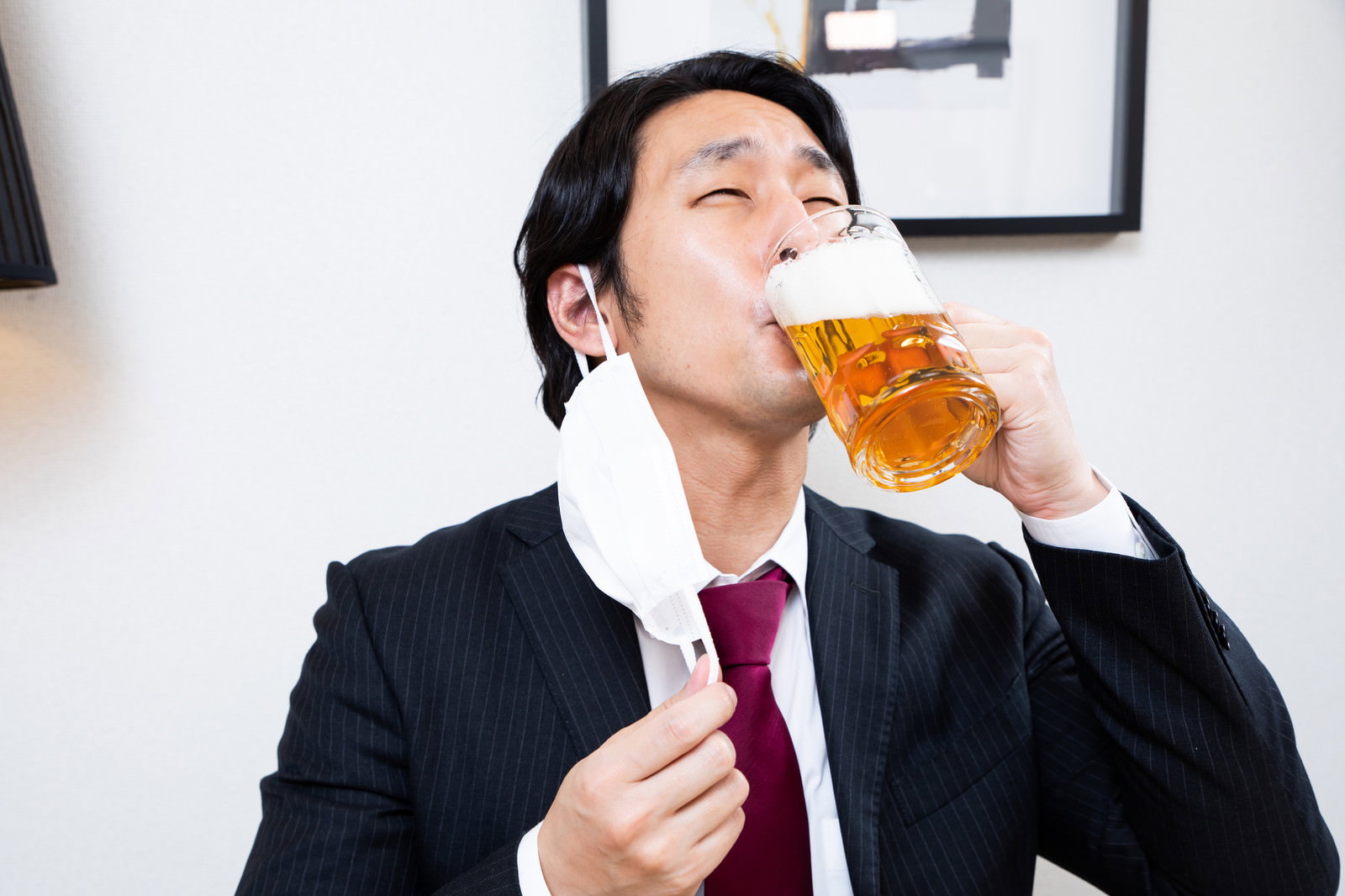 マスクを外してジョッキで生ビールを飲むスーツ姿の男性［モデル：大川竜弥］