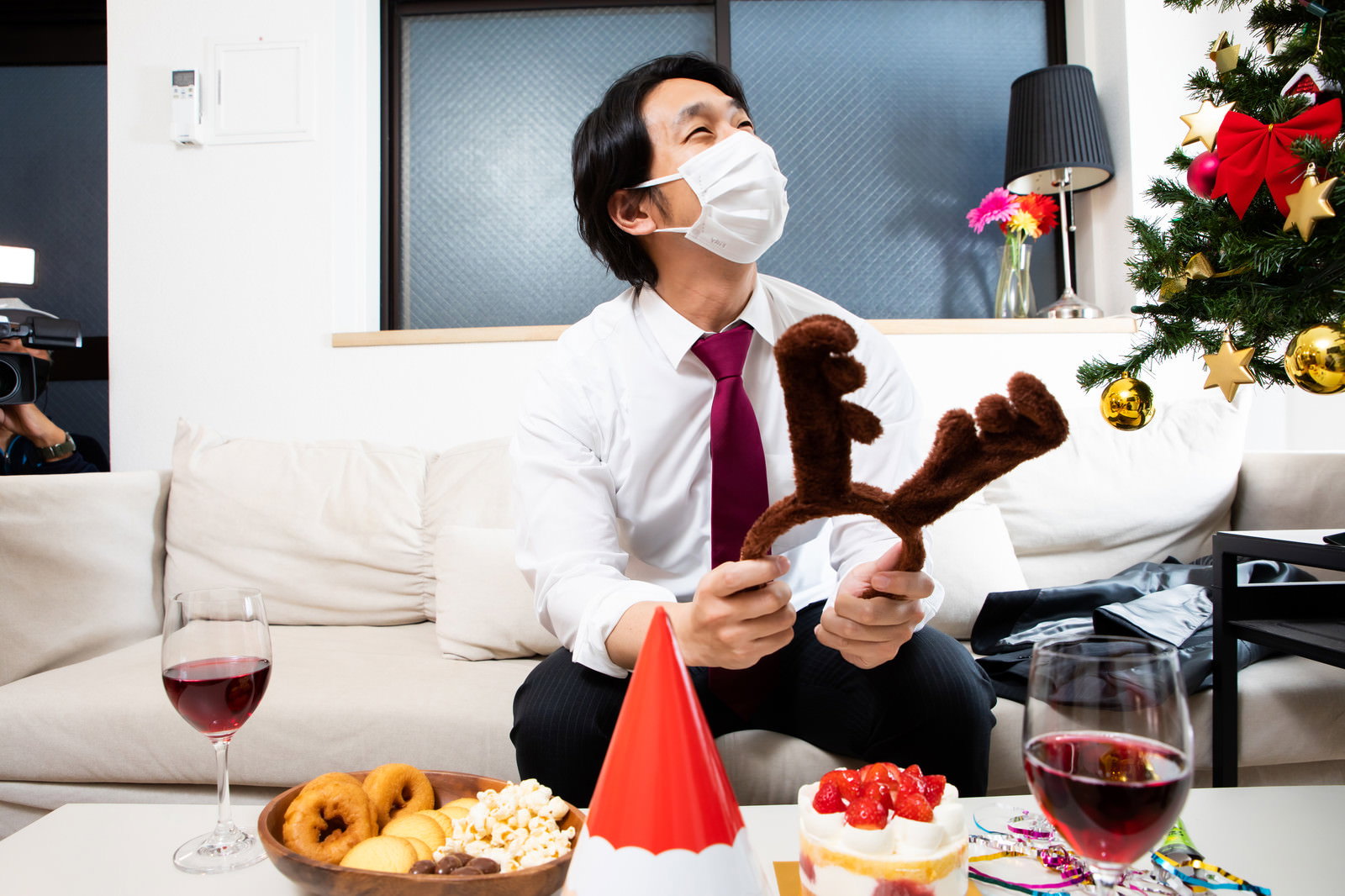 マスク姿でトナカイカチューシャを持ち、ソファーで赤ワインを飲む男性のクリスマス風景［モデル：大川竜弥］