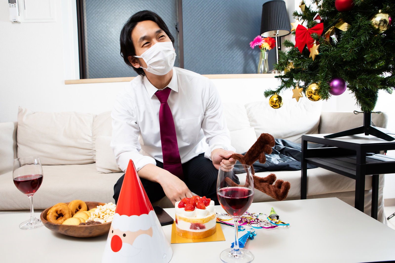 白いマスクを着用したスーツ姿の男性がソファーに座りクリスマス料理とワインが並ぶテーブルの前にいる［モデル：大川竜弥］