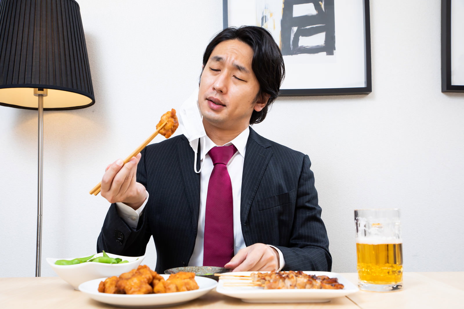 唐揚げを箸で掴んで食べるスーツ姿の会社員男性と生ビールのジョッキ［モデル：大川竜弥］
