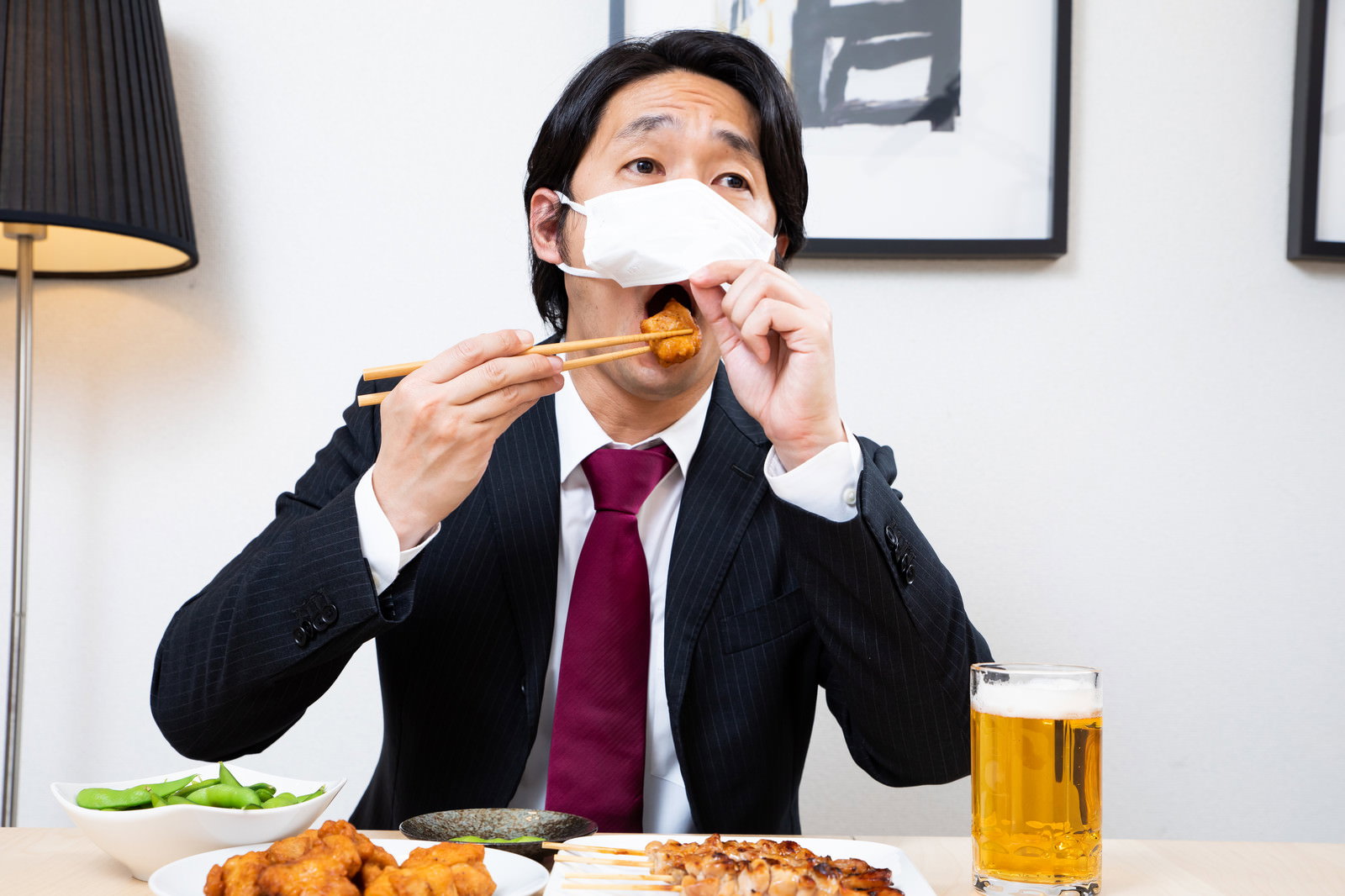 マスクを下げて唐揚げを口に入れるスーツ姿の会社員［モデル：大川竜弥］