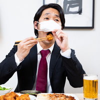 ずらしたマスクから唐揚げを口に入れる会社員男性の写真