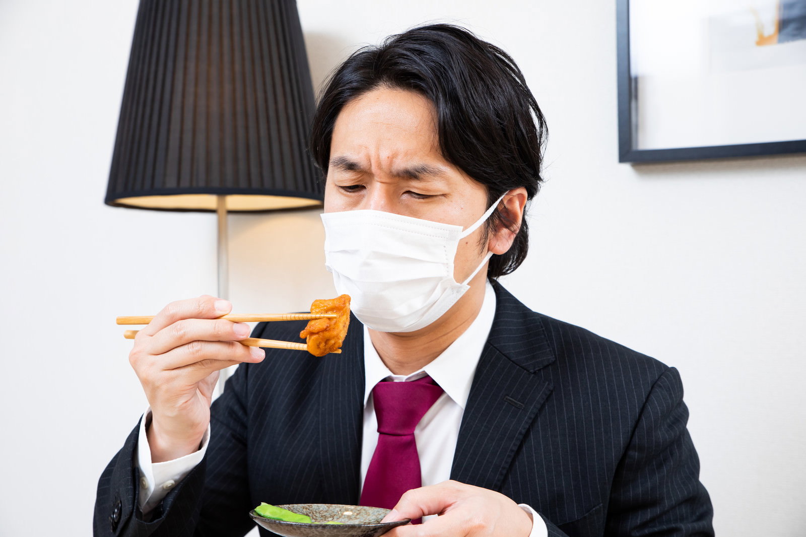 箸で唐揚げを摘み、マスクを着用したまま食べ物を見つめる会社員男性［モデル：大川竜弥］