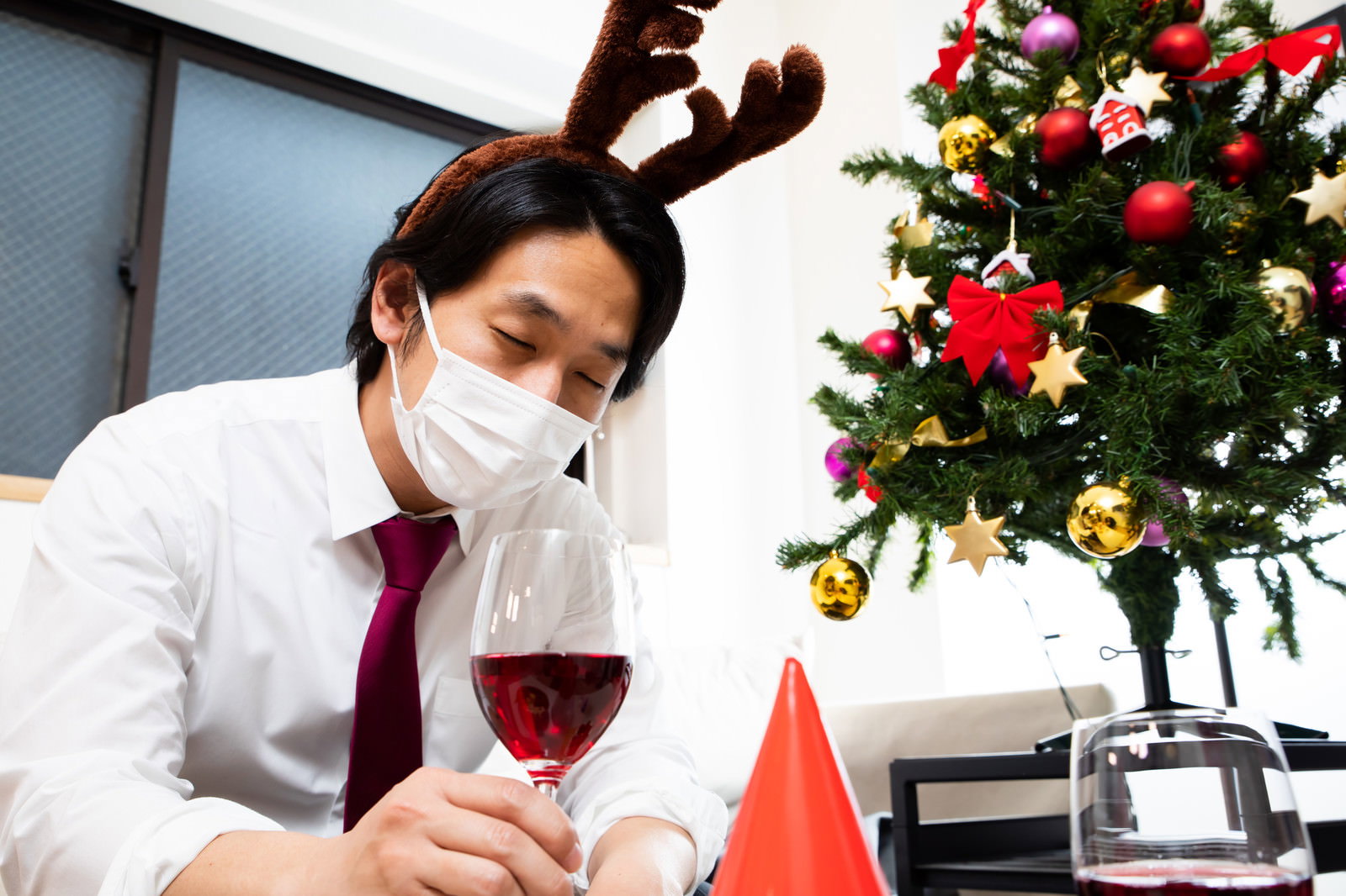 赤ワインを手に眠るマスク姿の男性。トナカイの角、紫ネクタイ、背景のクリスマスツリーと装飾品［モデル：大川竜弥］