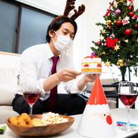 トナカイの角をつけてクリスマスケーキを見つめるマスク着用の会社員の写真