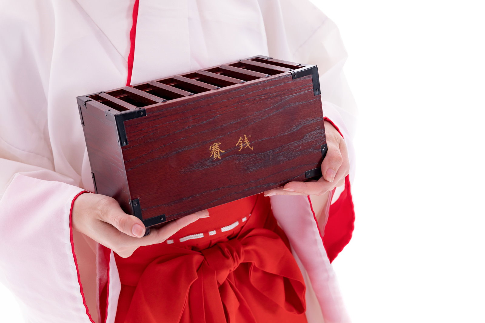 白い上衣と赤い袴を着た巫女が赤い紐の付いた木製の携帯賽銭箱を両手で持っている様子［モデル：緋真煉］