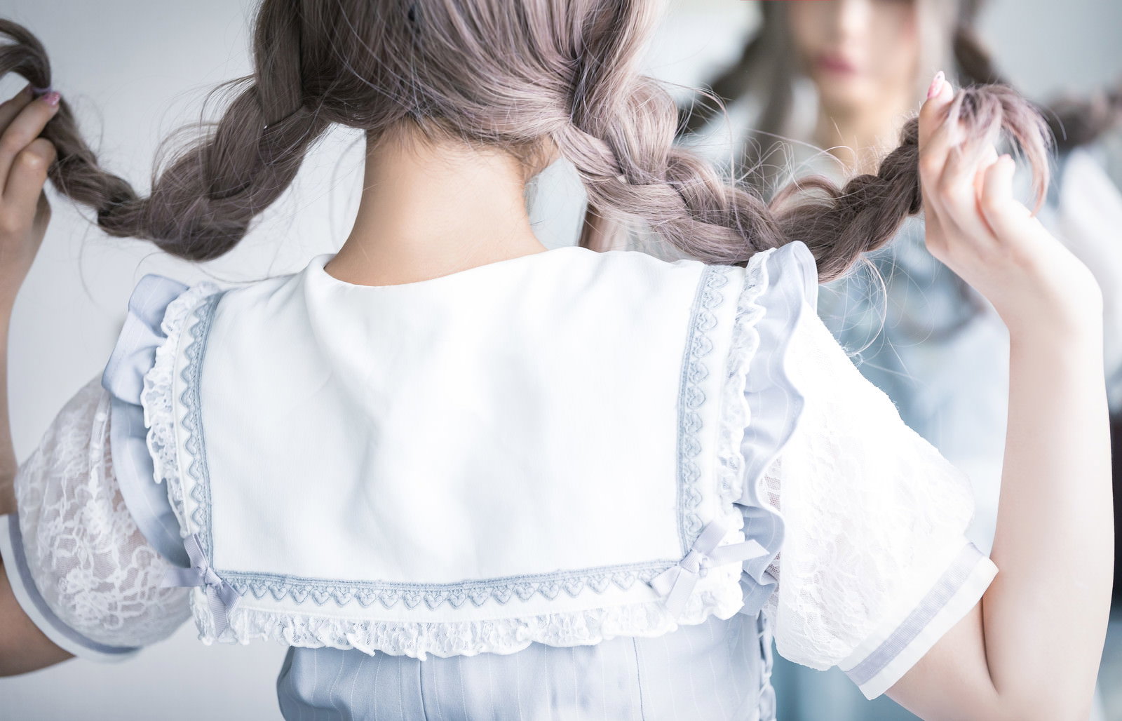 白いエプロンドレス風の衣装を着た女性が鏡の前で三編みヘアを両手で持つ後ろ姿［モデル：石投げて美奈代］