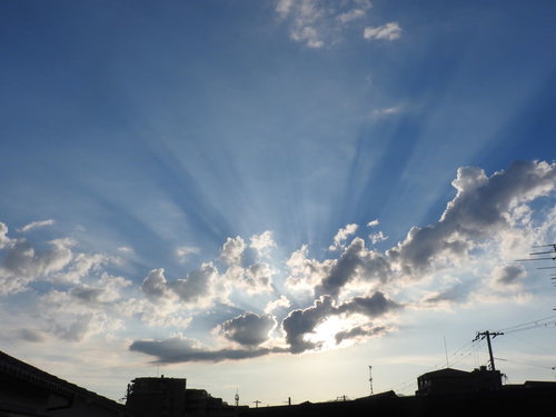 空にかかる夕陽の光芒と建物のシルエット、雲間の薄明光線