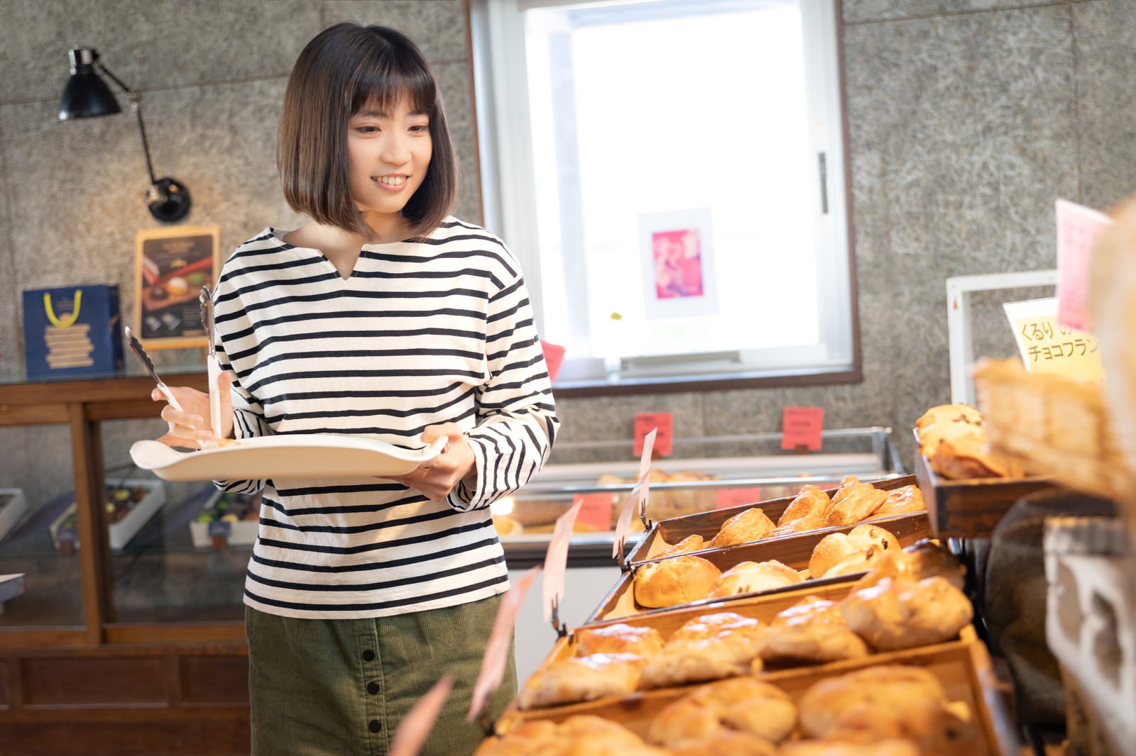 パン屋の店内でボーダーシャツの女性が菓子パンを選んでいる［モデル：塩田みう］