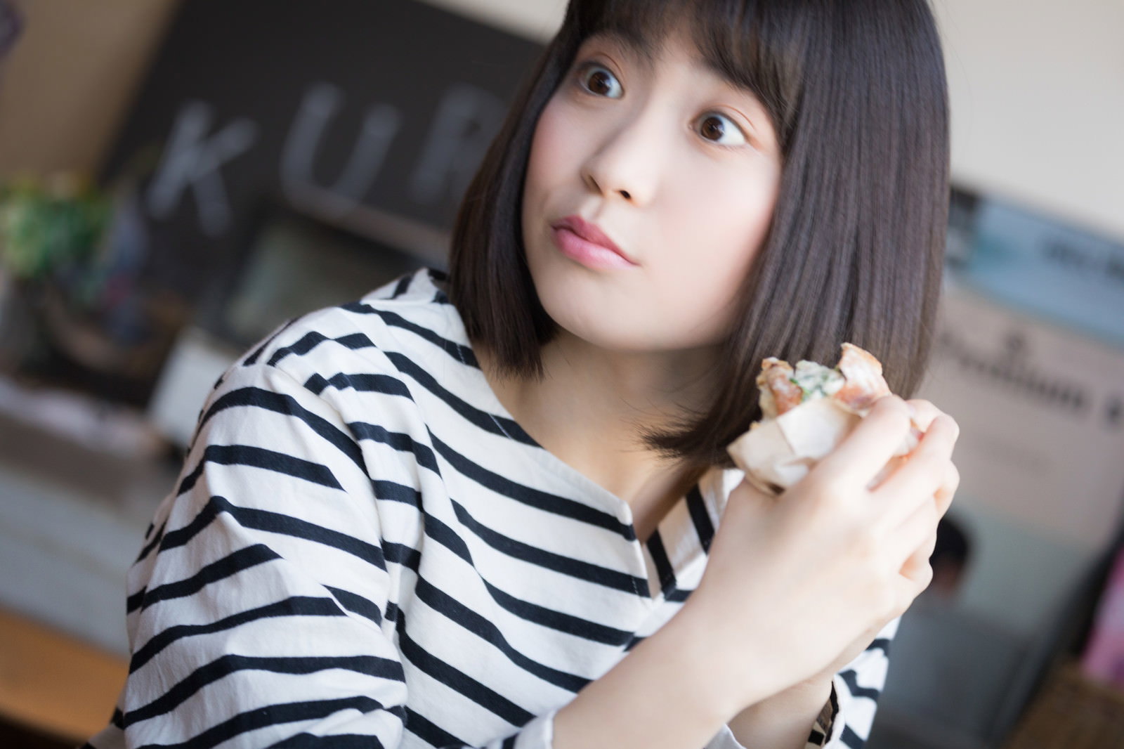 フードコートでボーダーシャツを着た女性がパンを食べながらスマホを操作している［モデル：塩田みう］