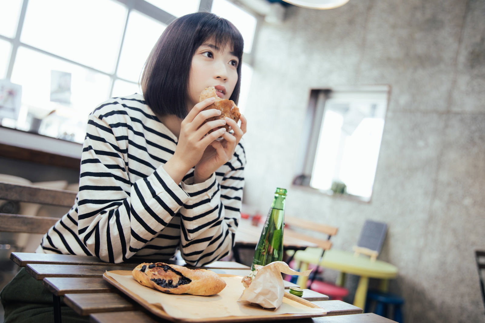 フードコートのテーブルで菓子パンを食べるボーダーシャツの女性［モデル：塩田みう］