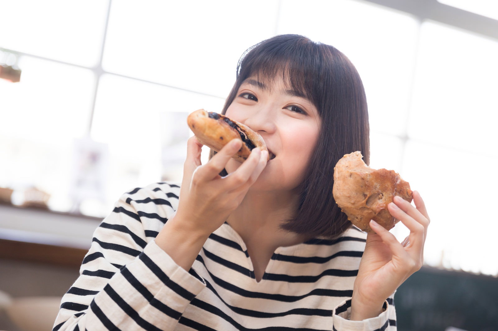 ボーダーシャツの女性が両手にパンを持って笑顔で食べている［モデル：塩田みう］