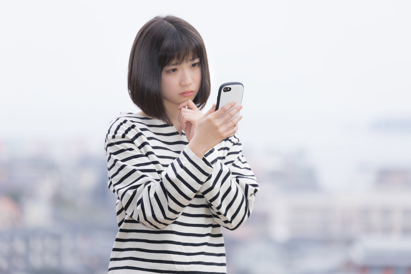 スマートフォンを見つめて困った表情を浮かべるボーダーシャツの女性［モデル：塩田みう］