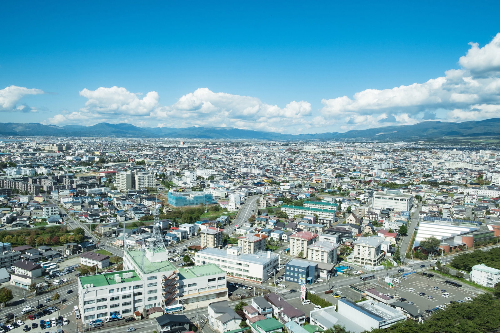 青空の下、高台から俯瞰で見た函館市街地に住宅やビルが広がり奥に山並みが連なる風景