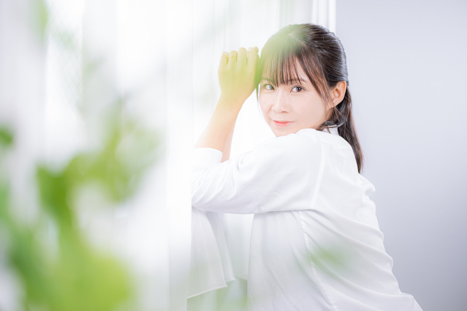 窓に寄り掛かり髪に手をやる白シャツの女性［モデル：本坊実結］