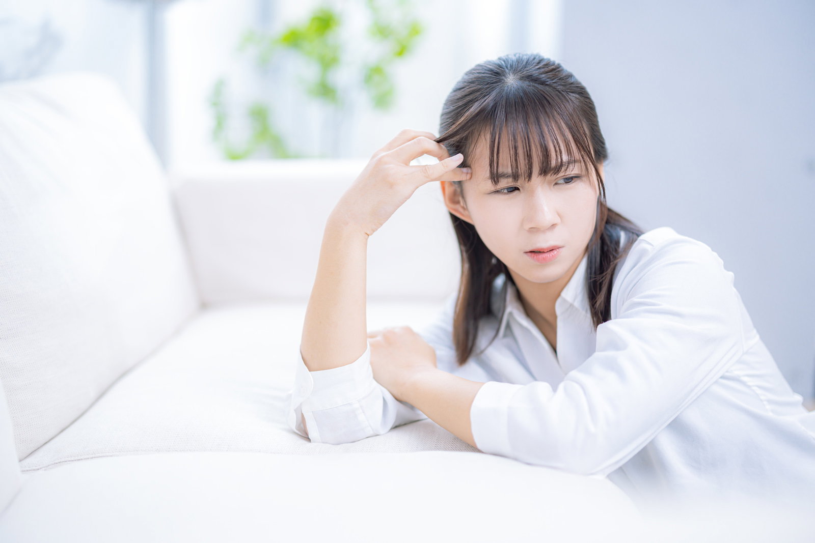ソファーで頭痛に苦しむ女性が手を頭に当てている［モデル：本坊実結］