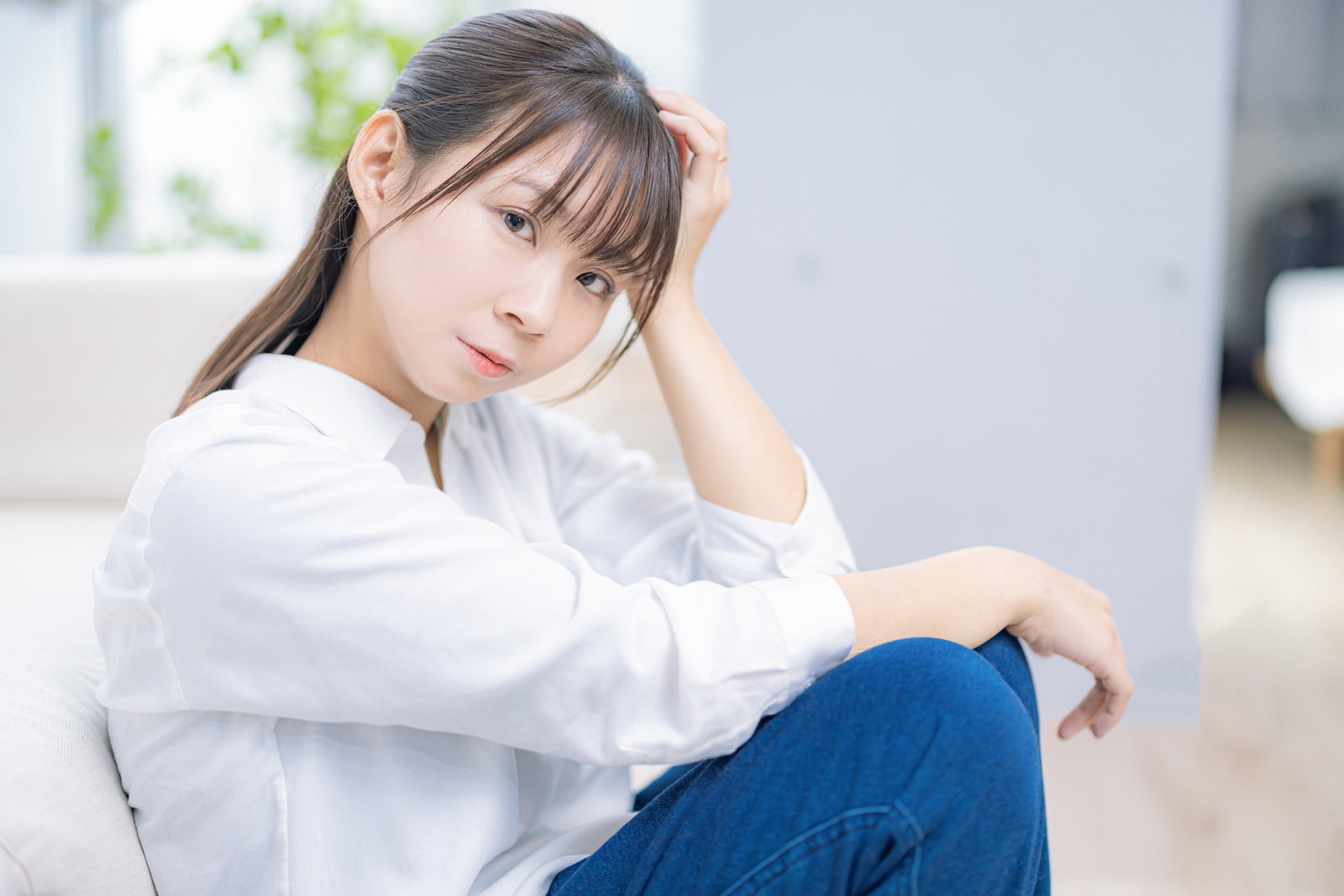 白いシャツを着た女性がソファーにもたれかかり、髪に手をやりながら思い悩んでいる［モデル：本坊実結］