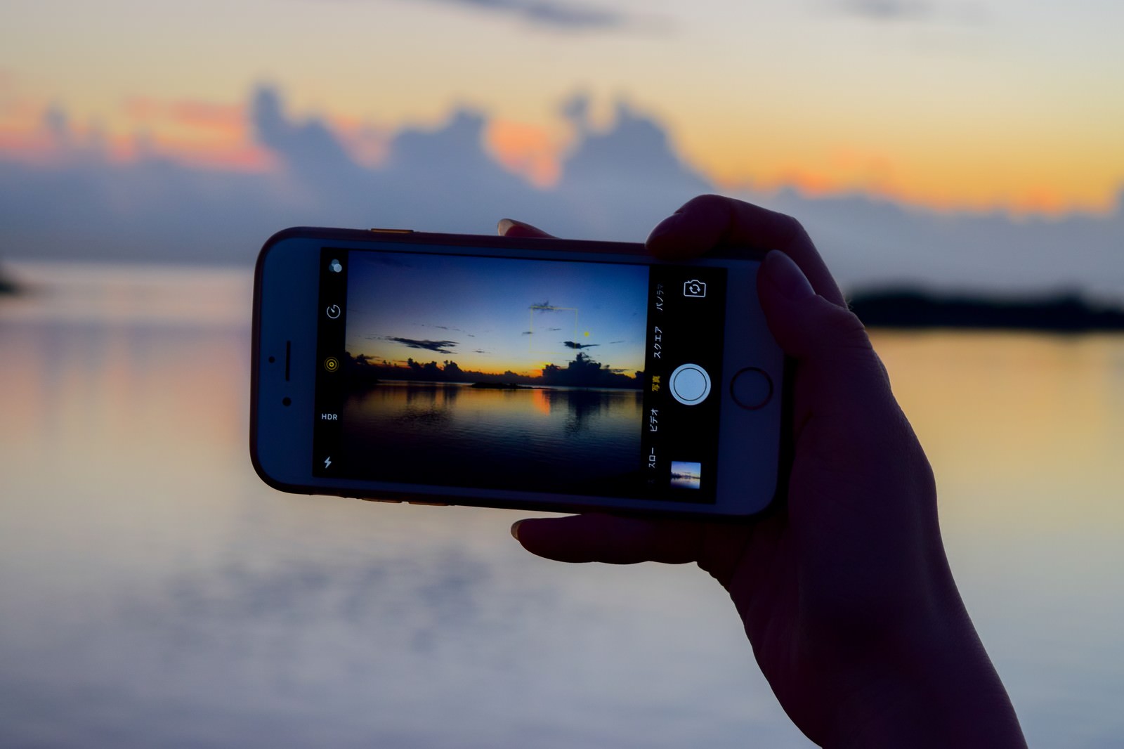 夕焼けの海をスマートフォンで撮影している手のシルエット