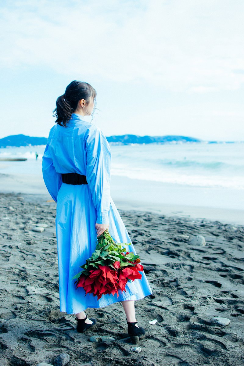 波打ち際の砂浜で赤いバラの花束を手に持つ青い服の女性