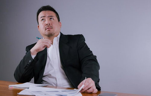 顎に手を当てて斜め上を見上げながら考え込む黒いスーツのビジネスマン