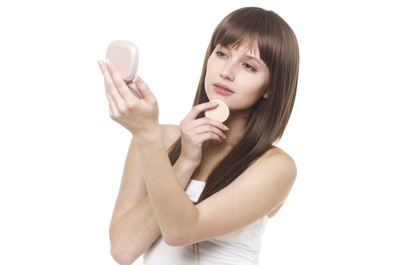 ピンクのコンパクトミラーを見ながらパフで化粧直しをする若い女性［モデル：モデルファクトリー］