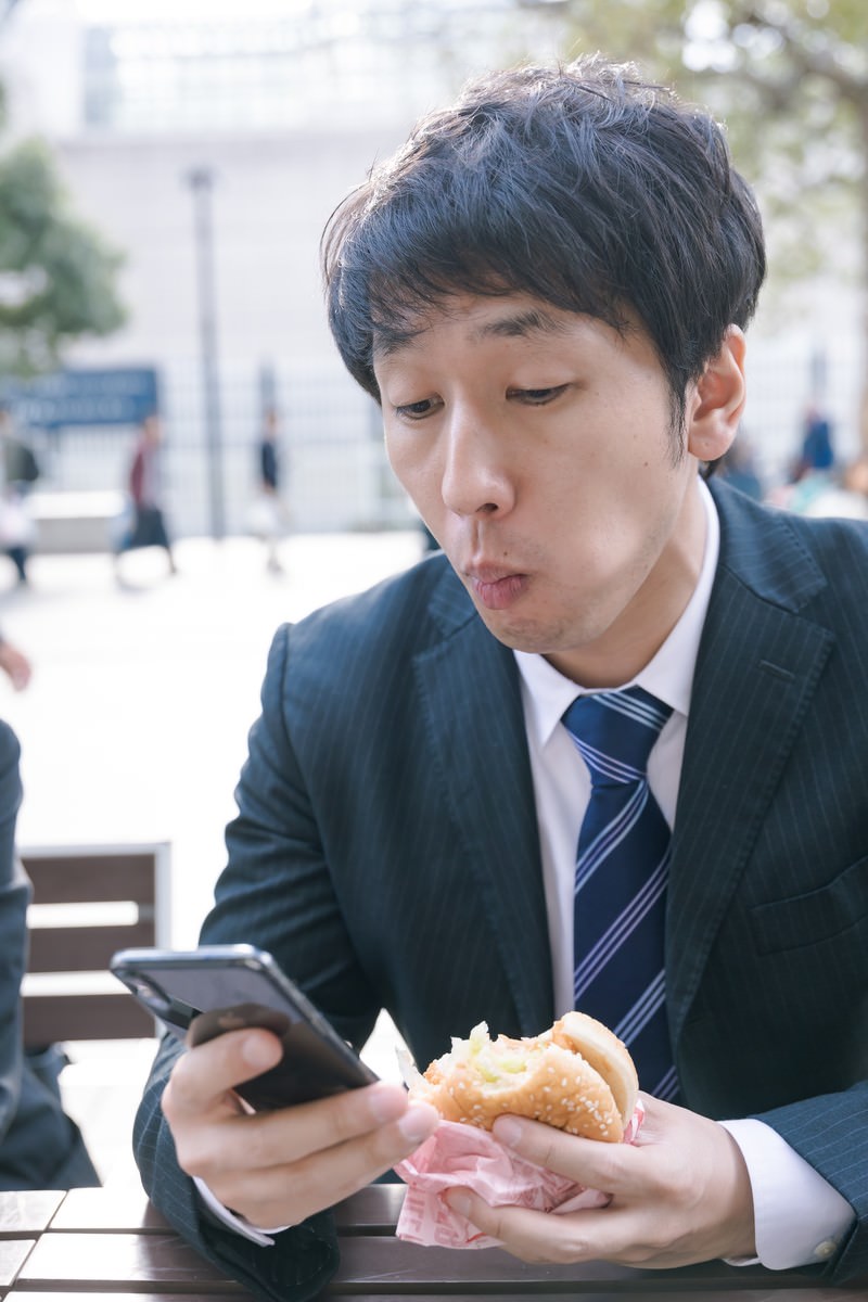 黒いスーツを着た男性がハンバーガーを手に持ちながらスマートフォンを操作している様子［モデル：大川竜弥］