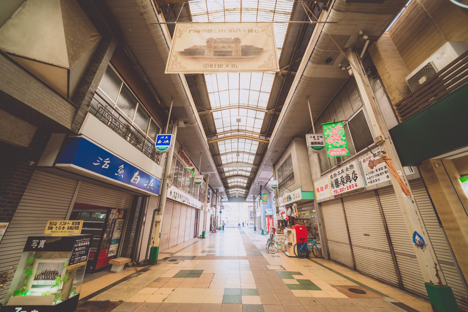 シャッターが閉じたアーケード商店街の通路。ガラス天井から光が差し込み、両側に店舗と懐かしい看板が見える