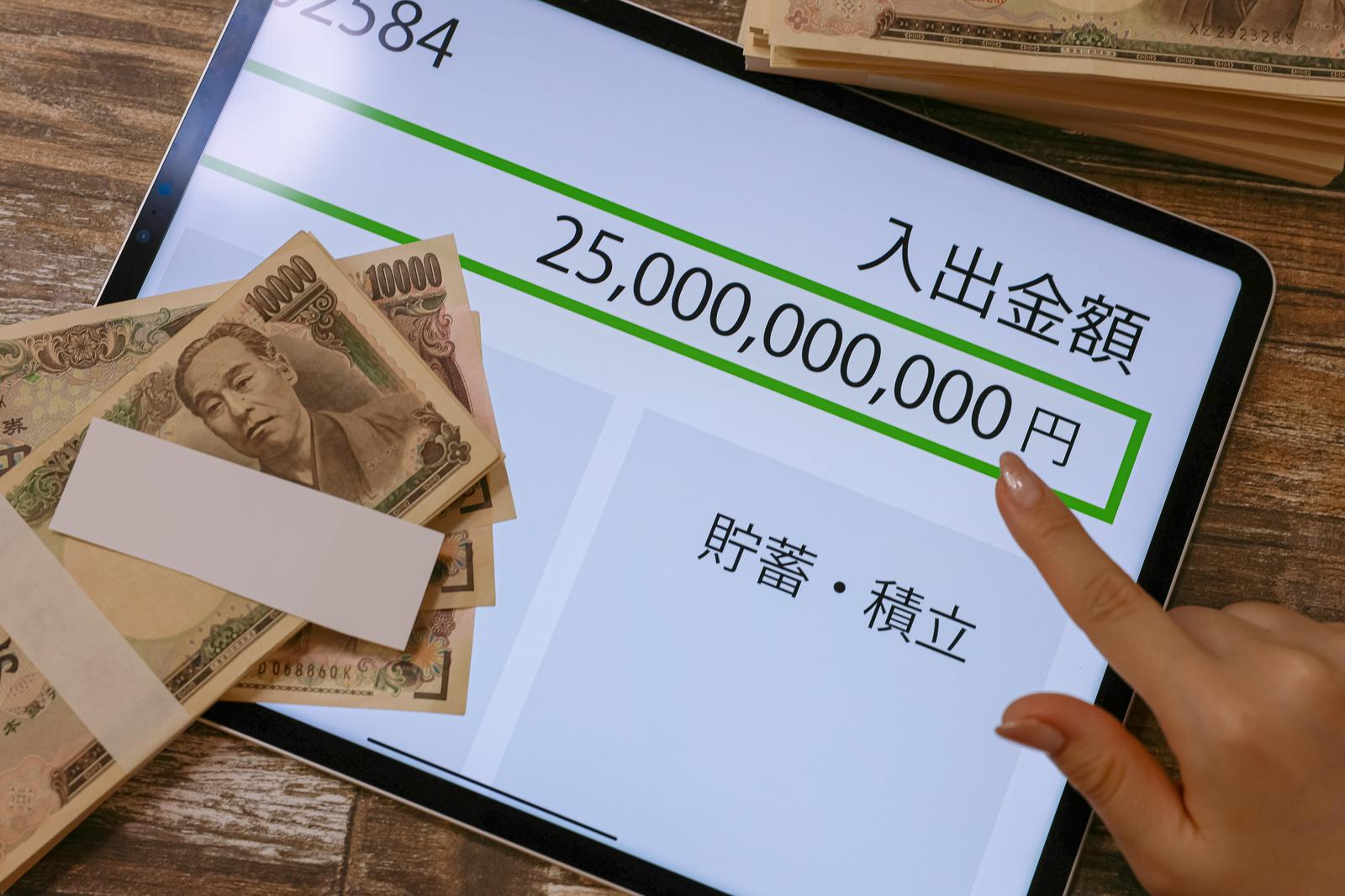 タブレットの画面に表示された高額な入出金額と札束を指差す手元