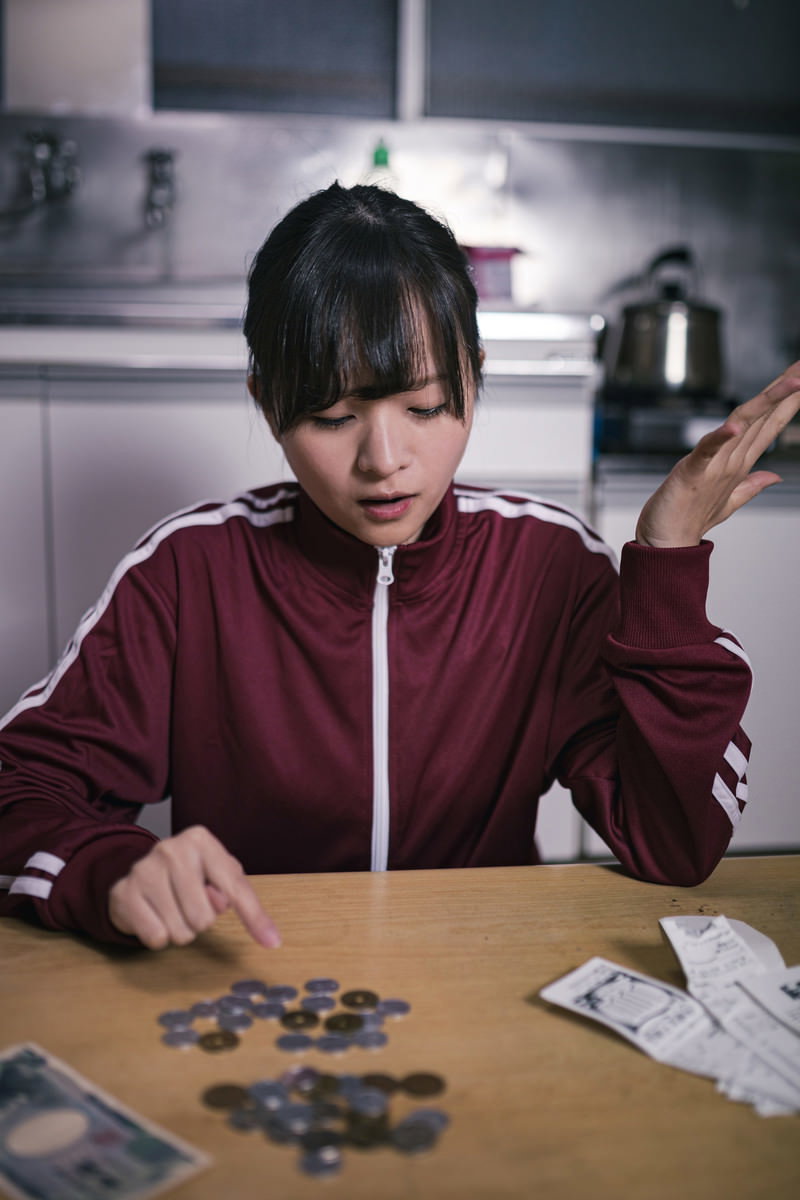 テーブルでレシートと小銭を見比べて困っている赤いジャケットの女性［モデル：千歳］