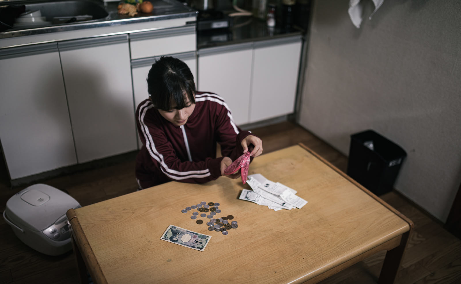室内のテーブルに座り込み、小銭を前に頭を抱える女性［モデル：千歳］