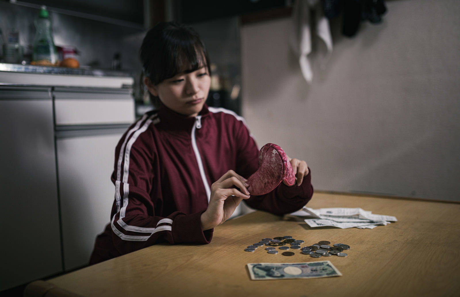 ワンルームのテーブルで金欠に困っている赤いジャージの女性［モデル：千歳］